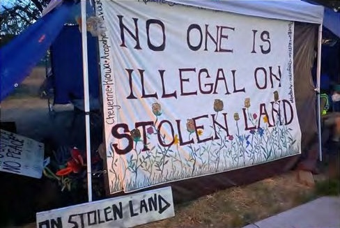 A quilt hangs from a canopy tent with flowers growing behind large red text that reads, "NO ONE IS ILLEGAL ON STOLEN LAND". Smaller stitched text going up the left side of the quilt reads, "Cheyenne, Kiowa, Araphaoe, Navajo".