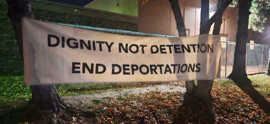 White banner hanging between two trees displays two lines of english text in black. First line reads: "Dignity Not Dentention". Second line reads: "End Deportations"