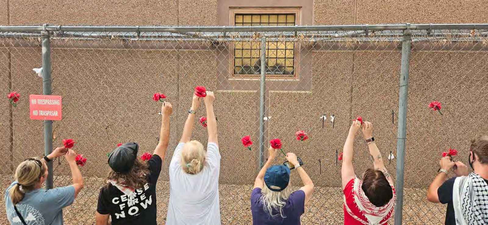 Six people stand on the outside of the detention facility with their arms reaching up to append paper roses to show solidarity with those detained. A red sign reads, 'No Trespassing No Traspasar'.